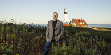 Boots2Roots Executive Director Randy Bell stands among tall grass with a charming lighthouse and water in the background.